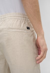 Nahaufnahme einer Hand, die neben beigen und weißen gestreiften Hosen mit einem elastischen Bund und einem kleinen schwarzen Markenlabel ruht.