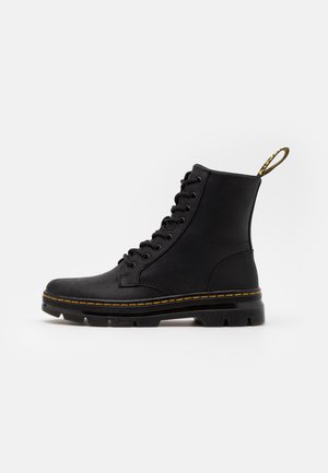 Botte haute en cuir noir avec coutures jaunes, lacets à l'avant, semelle robuste, et languette d'extraction à motif jaune et noir.