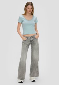 Jasnoniebieski prążkowany top z falbankowym dekoltem, zestawiony z szerokimi, wyblakłymi szarymi dżinsami. Modelka stoi pewnie, z rękami w kieszeniach.