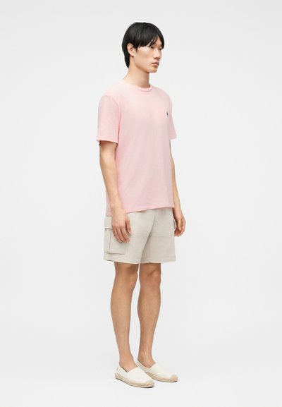 Άνδρας όρθιος που φοράει ανοιχτό ροζ μπλουζάκι, μπεζ σορτς cargo και λευκά slip-on εσπαντρίγιες, μπροστά σε απλό λευκό φόντο.