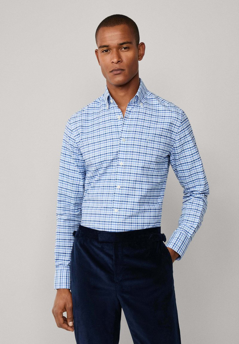 Man in een lichtblauw en wit geruit overhemd met een knoop, in combinatie met donkere marineblauwe broek, staand tegen een effen lichtgrijze achtergrond.