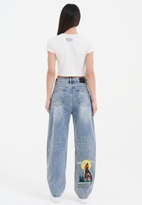Hvid cropped t-shirt med sort grafik på ryggen, parret med løse lyseblå denimjeans med et ulvedesign og teksten "Born to Be Wild".