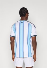 Balta sporta krekls ar vertikālām gaismas zilām svītrām, īsām piedurknēm un melniem akcentiem; uz kakla ir logs un teksts "1893".