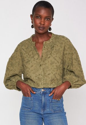 Woman wearing an olive green textured button-up blouse with balloon sleeves and high-waisted blue jeans, hands in front pockets.