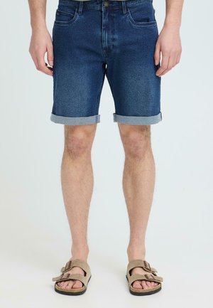 Homme portant un short en denim foncé retroussé et des sandales beiges à boucle, debout devant un fond clair uni.