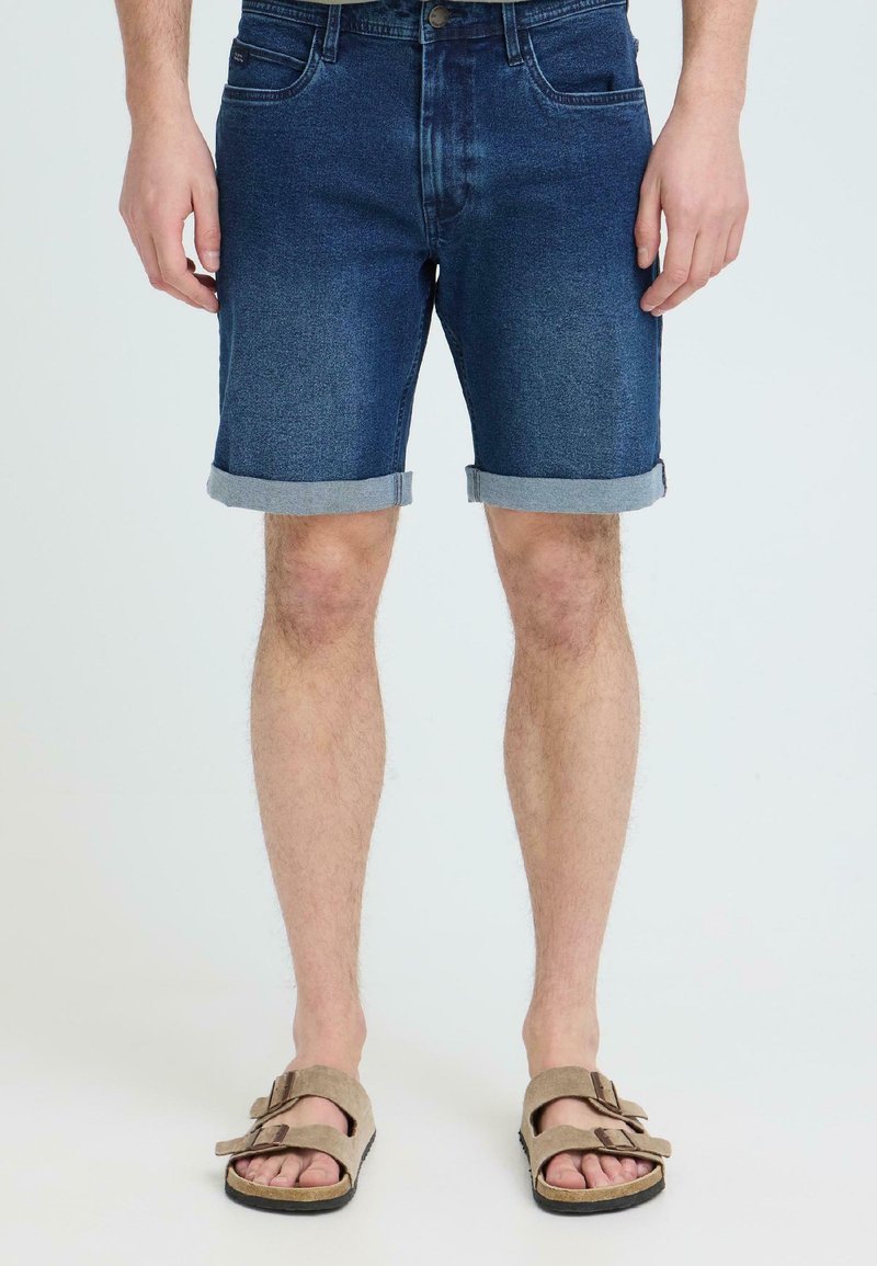 Mężczyzna w ciemnych, podwiniętych szortach z denimu i beżowych sandałach z klamrą, stojący na tle jasnego, jednolitego tła.