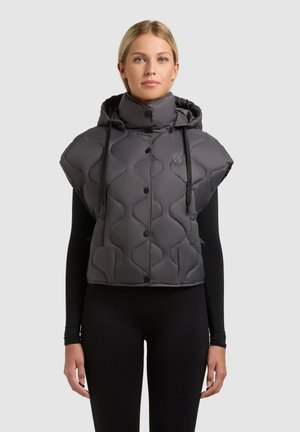 Kobieta ubrana w szarą pikowaną kamizelkę bez rękawów z kapturem, na czarnym długim rękawie i czarnych spodniach, na jednolitym tle.