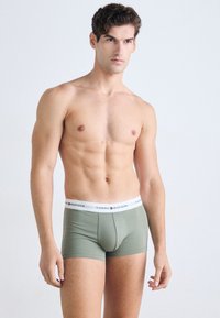 Mies, ktorý nemá na sebe tričko, s krátkymi kučeravými hnedými vlasmi, oblečený v zelených boxerkách Tommy Hilfiger, na jednoduchom svetlom pozadí.