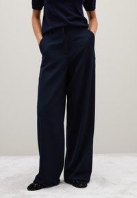 Marineblauwe wijde broek gemaakt van een soepele stof met zijzakken, gecombineerd met zwart paillettenschoenen, tegen een neutrale achtergrond.