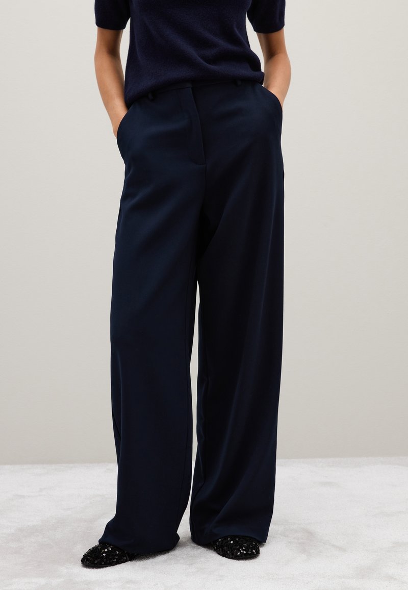 Marineblauwe wijde broek gemaakt van een soepele stof met zijzakken, gecombineerd met zwart paillettenschoenen, tegen een neutrale achtergrond.