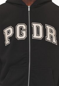 Černá mikina na zip vyrobená z látky, s bílými vyšívanými písmeny "PGDR" na hrudi a přední kapsou v pase.