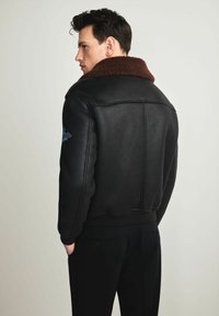 Czarna skórzana kurtka z brązowym kołnierzem z baranka, prążkowanymi mankietami i dopasowanym krojem. Posiada niebieską haftowaną naszywkę na ramieniu.