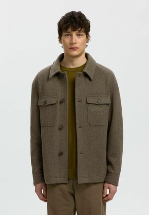Jeune homme portant une veste en laine marron à boutons sur une chemise vert olive, debout devant un fond clair uni.