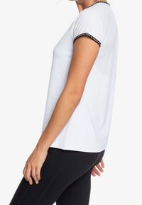 Vista lateral de una persona que lleva una camiseta corta blanca con ribetes negros y leggings negros sobre un fondo blanco liso.