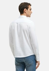 Weißes Langarmhemd mit Button-Down-Kragen, leicht glänzend und in lockerer Passform, kombiniert mit Jeans, von hinten betrachtet.