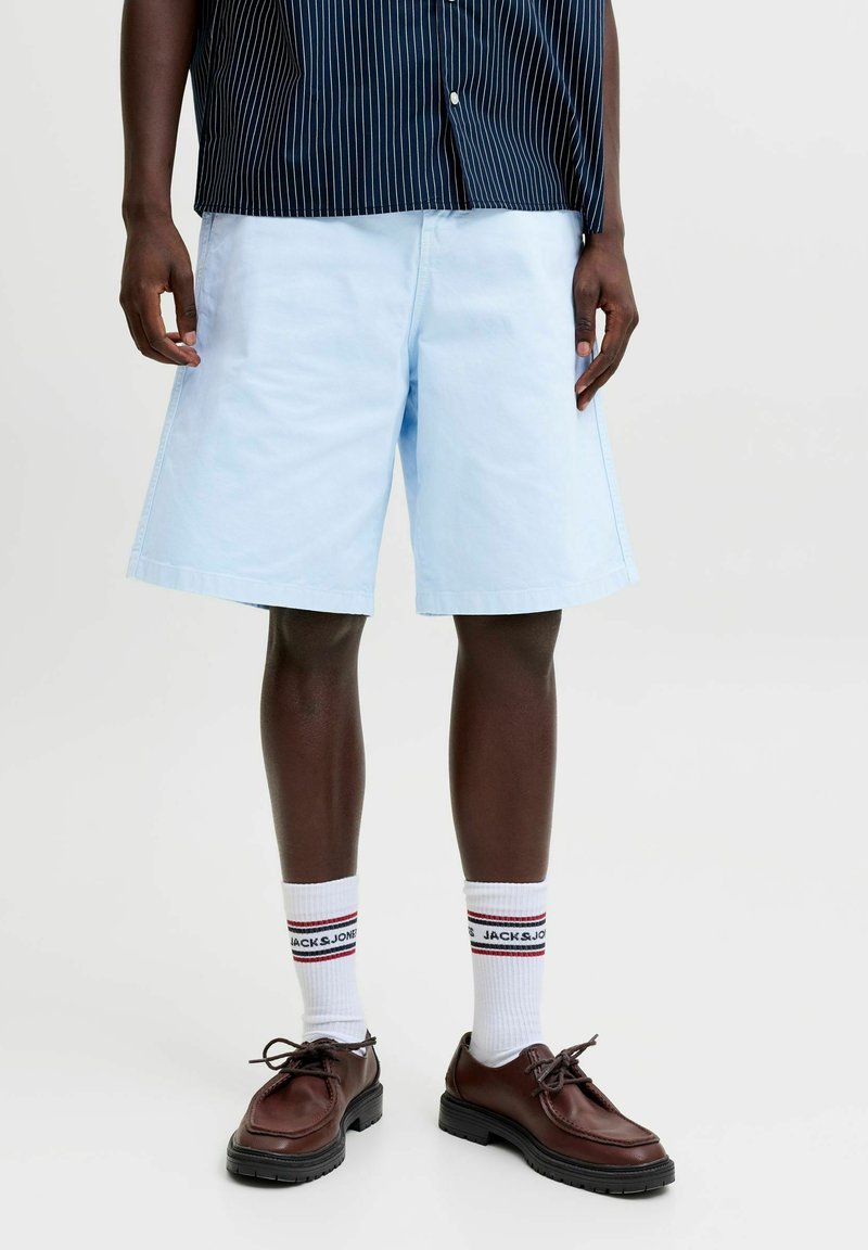 Homme portant un short bleu clair, des chaussettes blanches de marque, des chaussures en cuir marron et une chemise rayée bleu foncé, debout devant un fond uni.