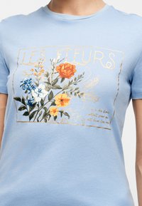T-shirt bleu avec un imprimé graphique floral comprenant des fleurs orange, jaune, blanche et bleue ainsi que le texte « LES FLEURS » en doré.