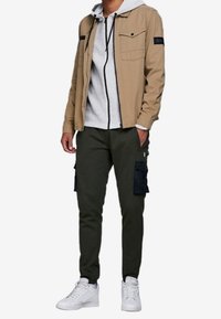 Tans jacket med bröstfickor över en grå huvtröja, parat med olivfärgade cargobyxor med sidofickor och vita sneakers.