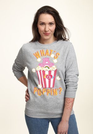 Szary sweter z kolorową grafiką niedźwiedzia w wiadrze popcornu, z napisem "CO SŁYCHAĆ?" w jasnych literach.