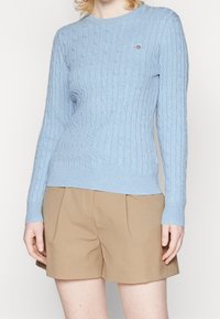 Jasnoniebieski dziany sweter z wzorem warkocza, okrągłym dekoltem i prążkowanymi mankietami. W połączeniu z beżowymi plisowanymi szortami. Brak widocznych logo.