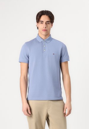 Tânăr în picioare, cu fața spre înainte, purtând un tricou polo bleu deschis cu mânecă scurtă și pantaloni bej pe un fundal alb simplu.