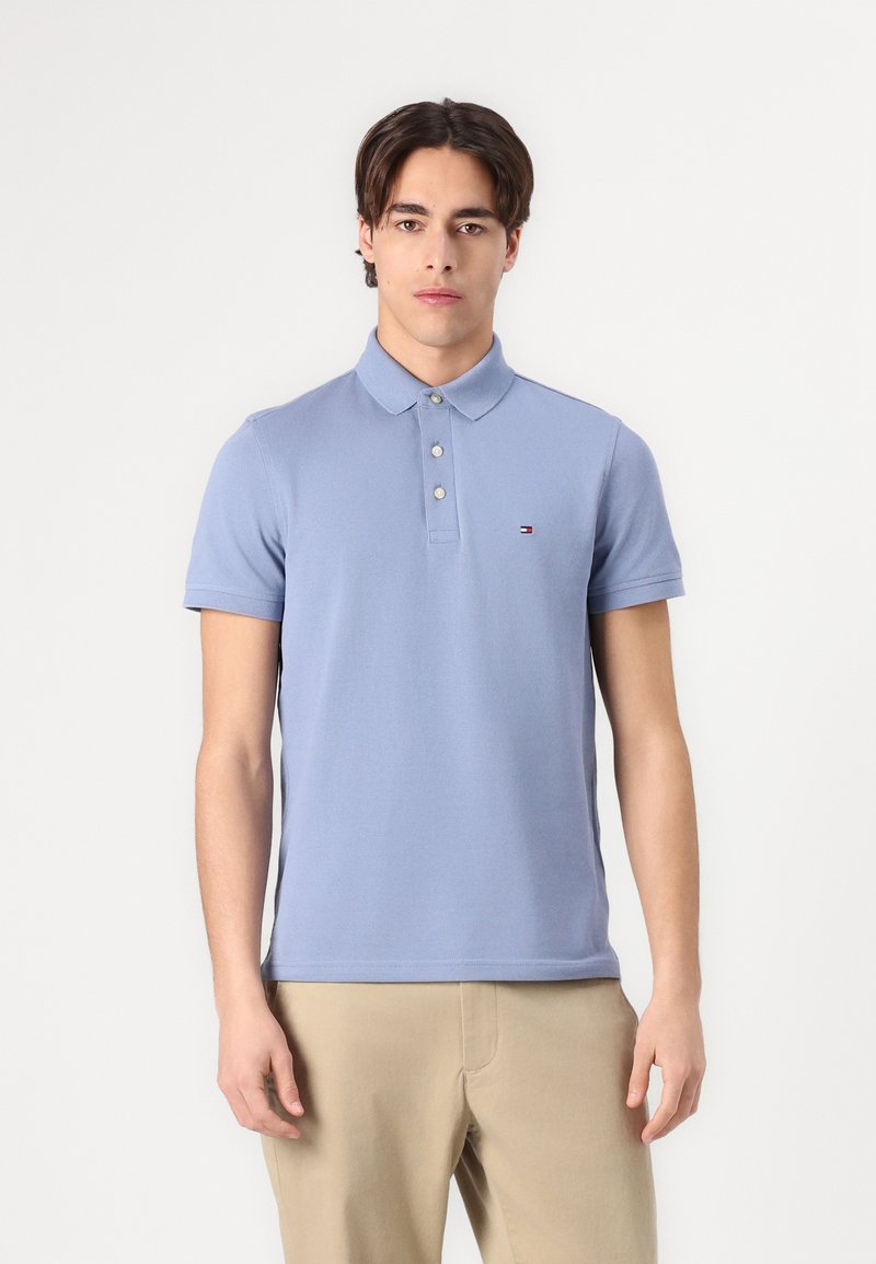 Ung mand, der står og kigger fremad iført en lyseblå kortærmet polo og beige bukser mod en ensfarvet hvid baggrund.