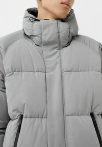 Světle šedá vycpaná bunda s vysokým límcem, obsahující zapínací boční kapsy a hladkou, elegantní texturu. Jednoduchý, moderní design.