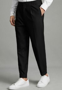 Pantalon noir en tissu lisse avec une taille élastique, un devant plissé et des jambes fuselées, associé à des baskets blanches.