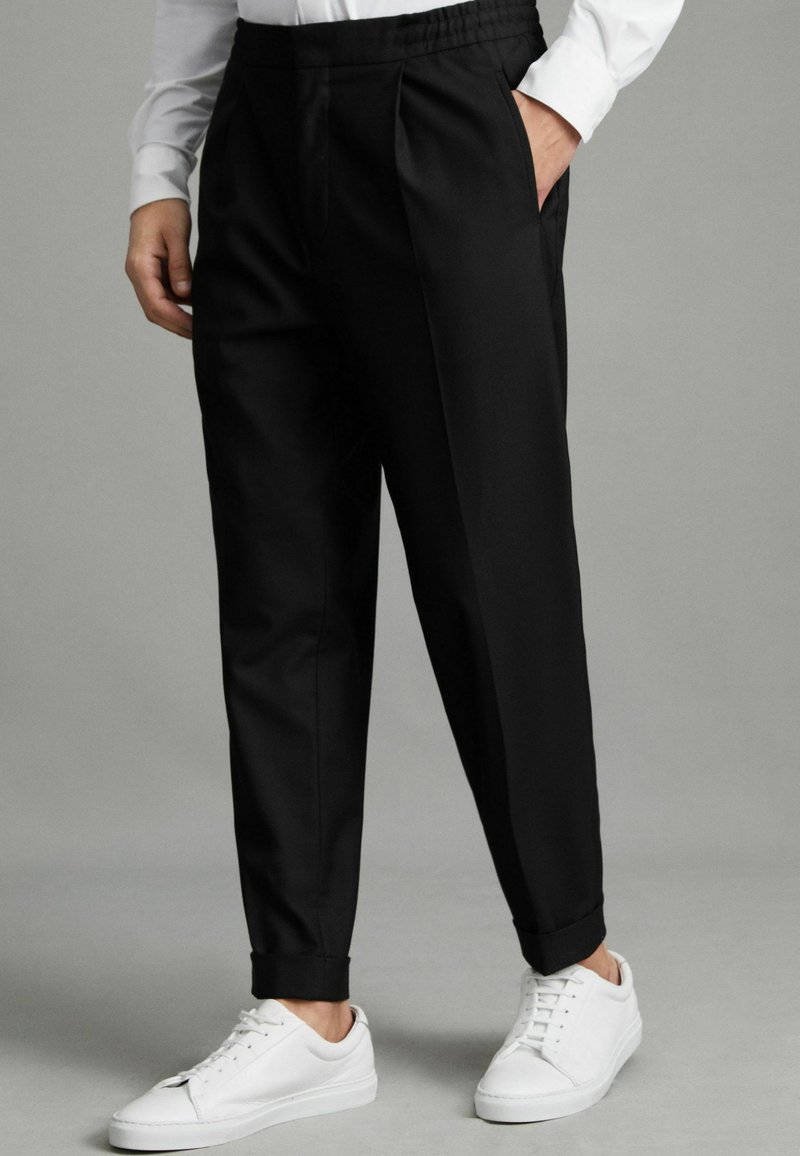 Pantalon noir en tissu lisse avec une taille élastique, un devant plissé et des jambes fuselées, associé à des baskets blanches.