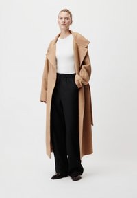 Cappotto di lana color cammello con rever ampi, indossato sopra una maglietta bianca e pantaloni neri. La texture è liscia, la silhouette è lunga e le scarpe scure completano l'outfit.