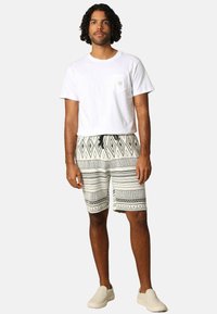 Witte katoenen t-shirt met zak, gecombineerd met beige en zwarte shorts met patronen en geometrische ontwerpen. Lichte instap-schoenen.