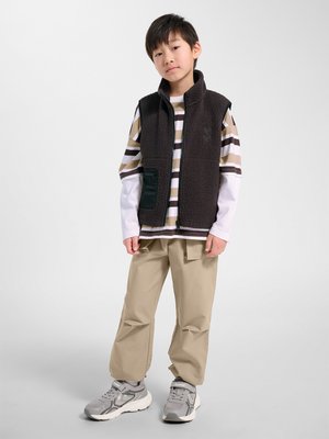 Junge steht, trägt eine schwarze Fleeceweste über einem braun-weiß gestreiften Langarmshirt, beigefarbene Hose und graue Sneakers vor einem grauen Hintergrund.