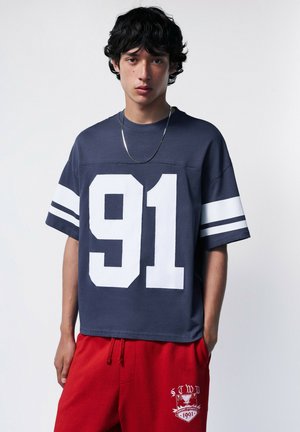 Junger Mann trägt ein marineblaues oversized T-Shirt mit weißer Zahl 91 und rote Jogginghose mit weißem Emblem, steht vor einfarbigem Hintergrund.
