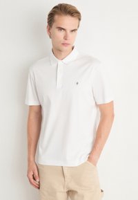 Camisa polo branca de manga curta, feita de algodão, com um colarinho clássico, placket com três botões e um pequeno logotipo no lado esquerdo do peito.