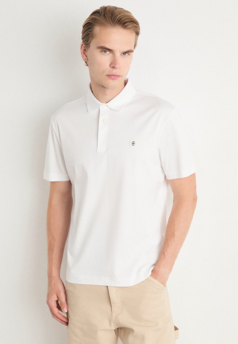 Camisa polo branca de manga curta, feita de algodão, com um colarinho clássico, placket com três botões e um pequeno logotipo no lado esquerdo do peito.