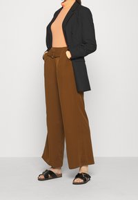 Schwarze Blazer, orangefarbener Rollkragenpullover, braune weite Hose mit gebundenem Taillenbund und schwarze Sandalen mit Schnalle. Glatte Stoffe, moderner Schnitt.