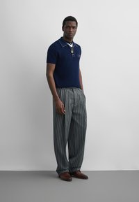 Homme portant un polo bleu marine à manches courtes, un pantalon large rayé, des chaussures marron et un pendentif en or, debout contre un mur gris uni.
