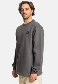 Sudadera gris de tela suave, con cuello redondo, puños acanalados, un pequeño parche de logo negro y un corte holgado.