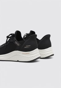 Schwarze Sportsneaker mit strukturiertem Obermaterial, Schnürverschluss, weißer konturierten Sohle und "Arch Comfort" Akzent an der Ferse.