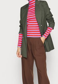 Person med en olivgrön blazer över en lila och röd randig ribbad polotröja, med bruna veckade byxor, hand i fickan.