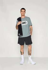 Sivá majica Nike z logotipom, črne kratke hlače, črn brisačo premeteno čez ramo, bele nogavice in bele teniske; tetovaže vidne na rokah.