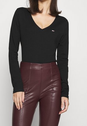 Femme portant un haut noir à manches longues avec un col en V et un pantalon en cuir marron foncé taille haute, debout devant un fond blanc.
