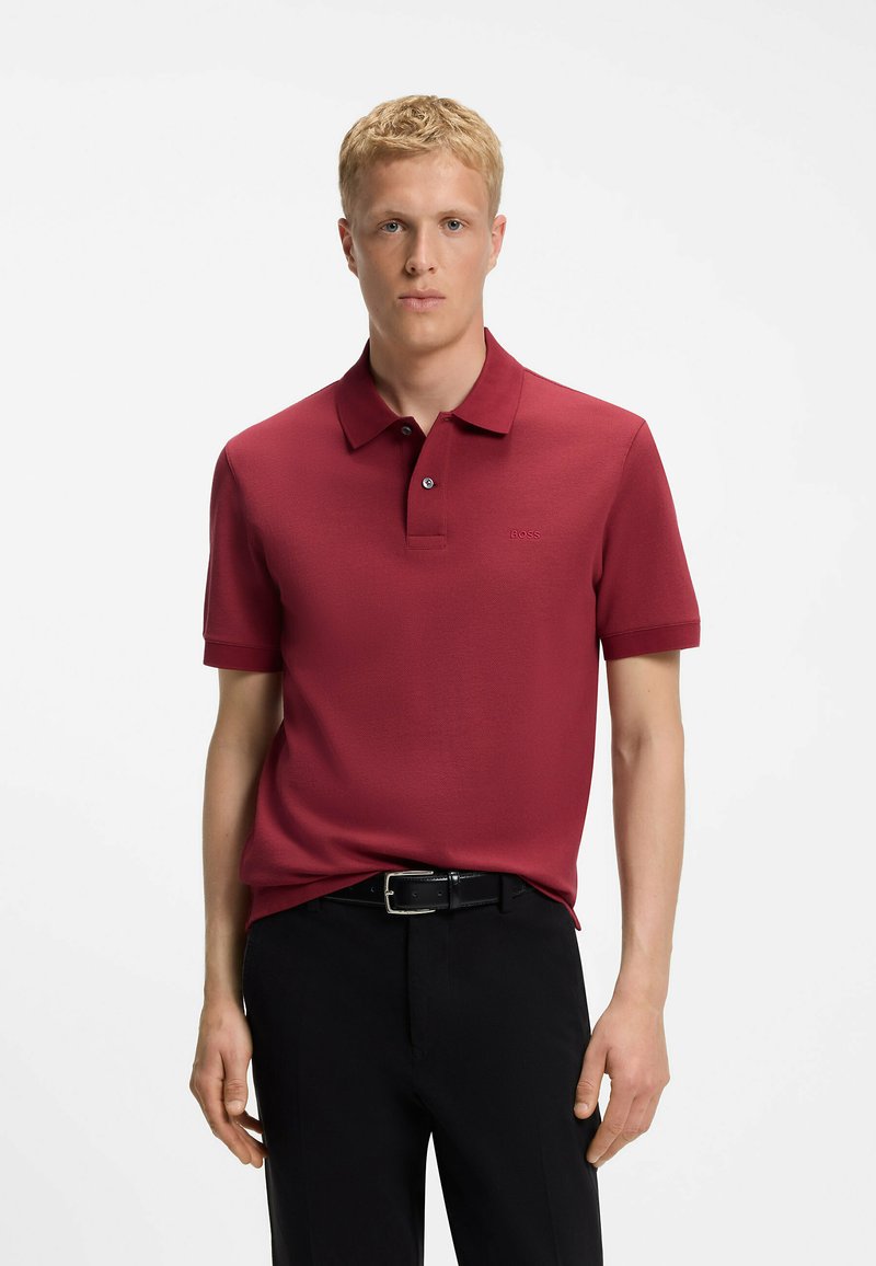 Jeune homme portant un polo rouge boutonné, rentré dans un pantalon noir avec une ceinture noire, debout devant un fond blanc uni.