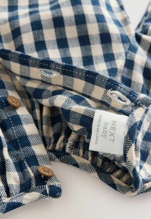 Mėlynos ir smėlio spalvos languota medžiaga su švelniu tekstūra, su rudais mygtukų akcentais ir etikete, nurodančia "NEXT baby, pagaminta Šri Lankoje."