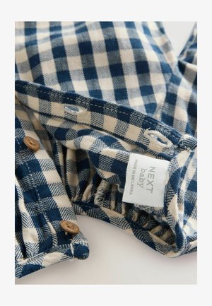 Sinine ja beež ruuduline kangas pehme tekstuuriga, pruunide nööbiga detailidega ja sildiga, millel on kiri "NEXT beebi, valmistatud Sri Lankas."