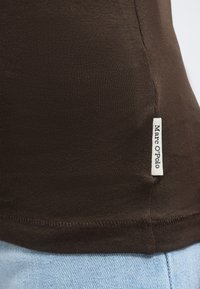 Κοντινό πλάνο σκούρας καφέ υφάσματος με μια λευκή ορθογώνια ετικέτα που γράφει "Marc O'Polo" ραμμένη στη ραφή στο πλάι πάνω από ανοιχτό μπλε τζιν.