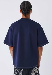 Navyblauwe oversized t-shirt met korte mouwen, gemaakt van gladde stof. Heeft korte zijsplitten en contrasterende stiksels langs de randen.