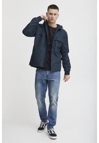 Marine jakke i lett materiale med hette, to frontlommer, elastiske mansjetter, båret over en svart skjorte, sammen med blå jeans og svarte sneakers.