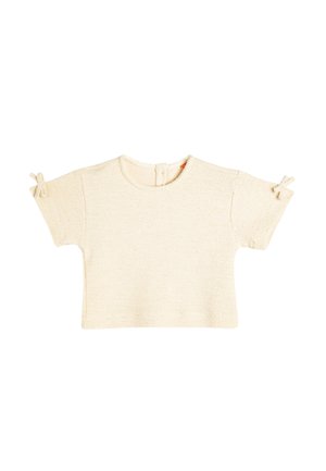 Camiseta de manga corta en color crema, fabricada en punto texturizado, con cuello redondo, cierre en la parte posterior y pequeños lazos en las mangas como detalle.