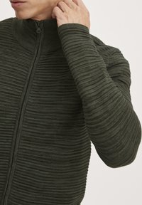 Grøn struktureret zip-up sweater med ribbet mønster, høj krave og tætsiddende ærmer, der viser et subtilt, vævet design.